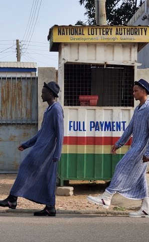 Two individuals are walking past a small kiosk with signage for the National Lottery Authority. The kiosk, decorated with a sign that reads 'Full Payment,' features yellow, green, red, and white colors. The individuals are dressed in matching long blue robes and hats, with one wearing black boots and the other white boots.